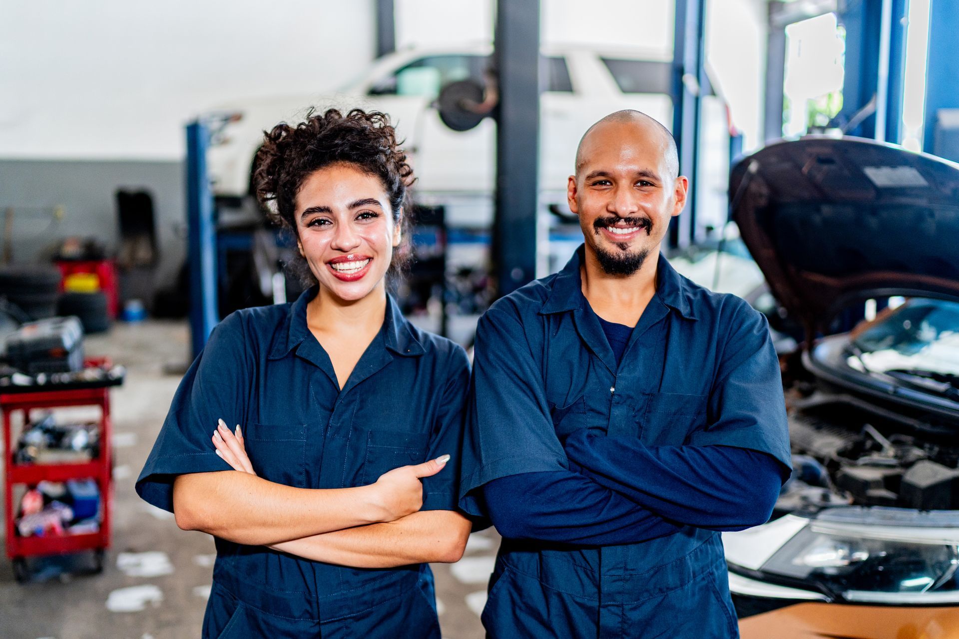 Two mechanics in blue jumpsuits smiling at the camera in a car repair shop.