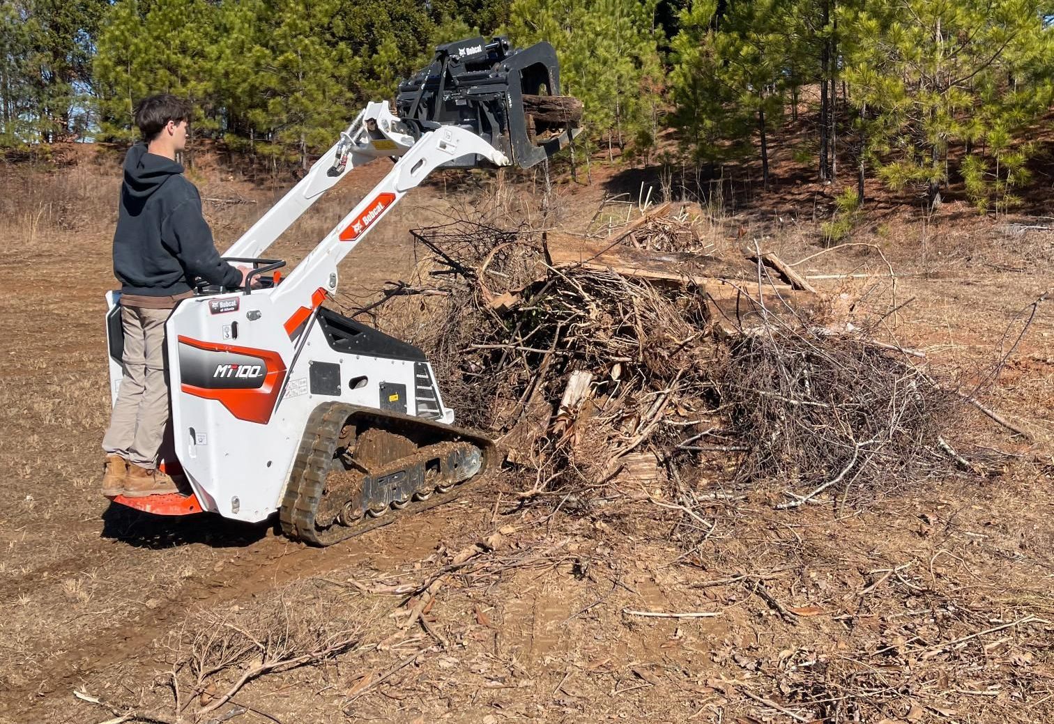 A person operating a Bobcat compact track loader, moving debris outdoors.