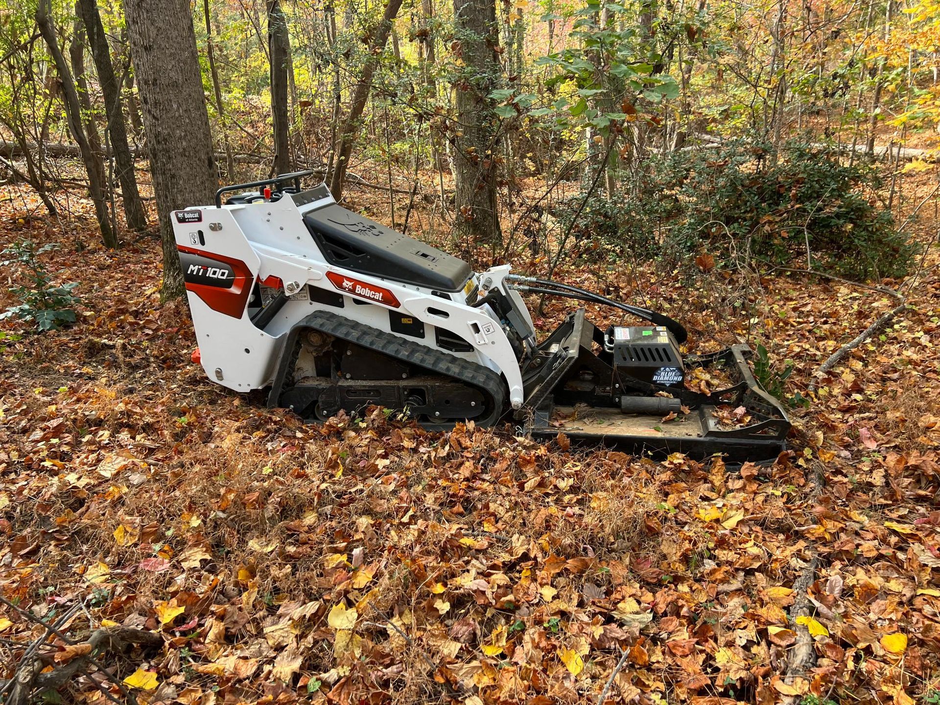 A white and red skid-steer loader with a mulching head in a wooded area covered in fallen leaves.