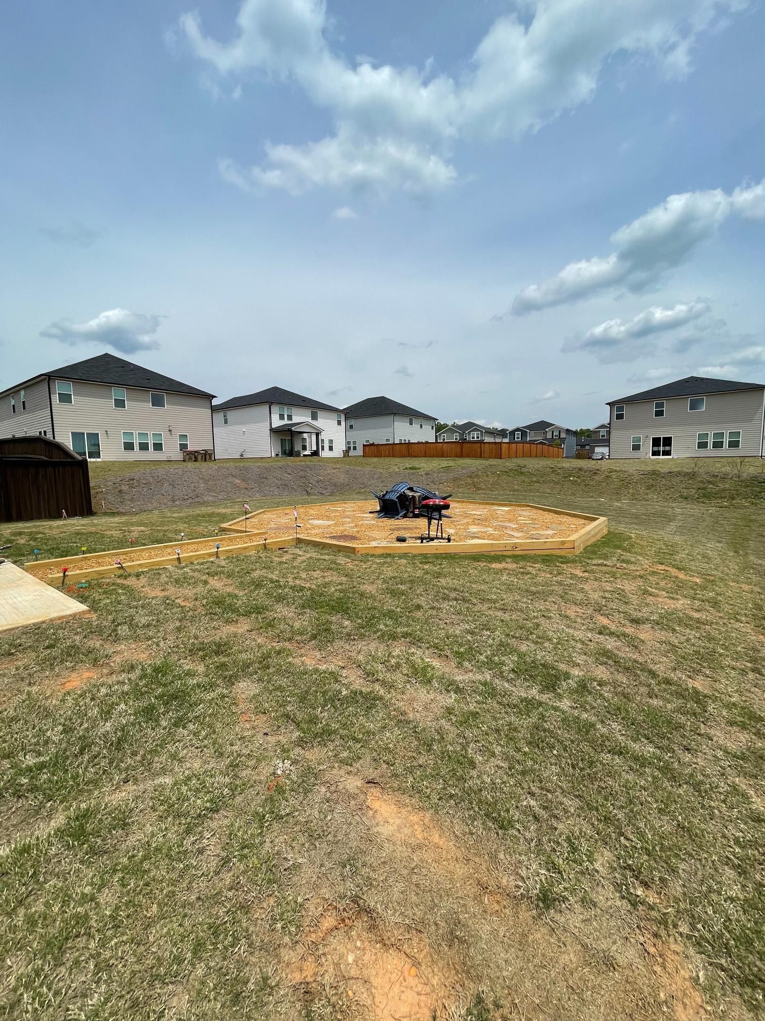 A large grassy field with houses in the background and a fire pit in the middle.