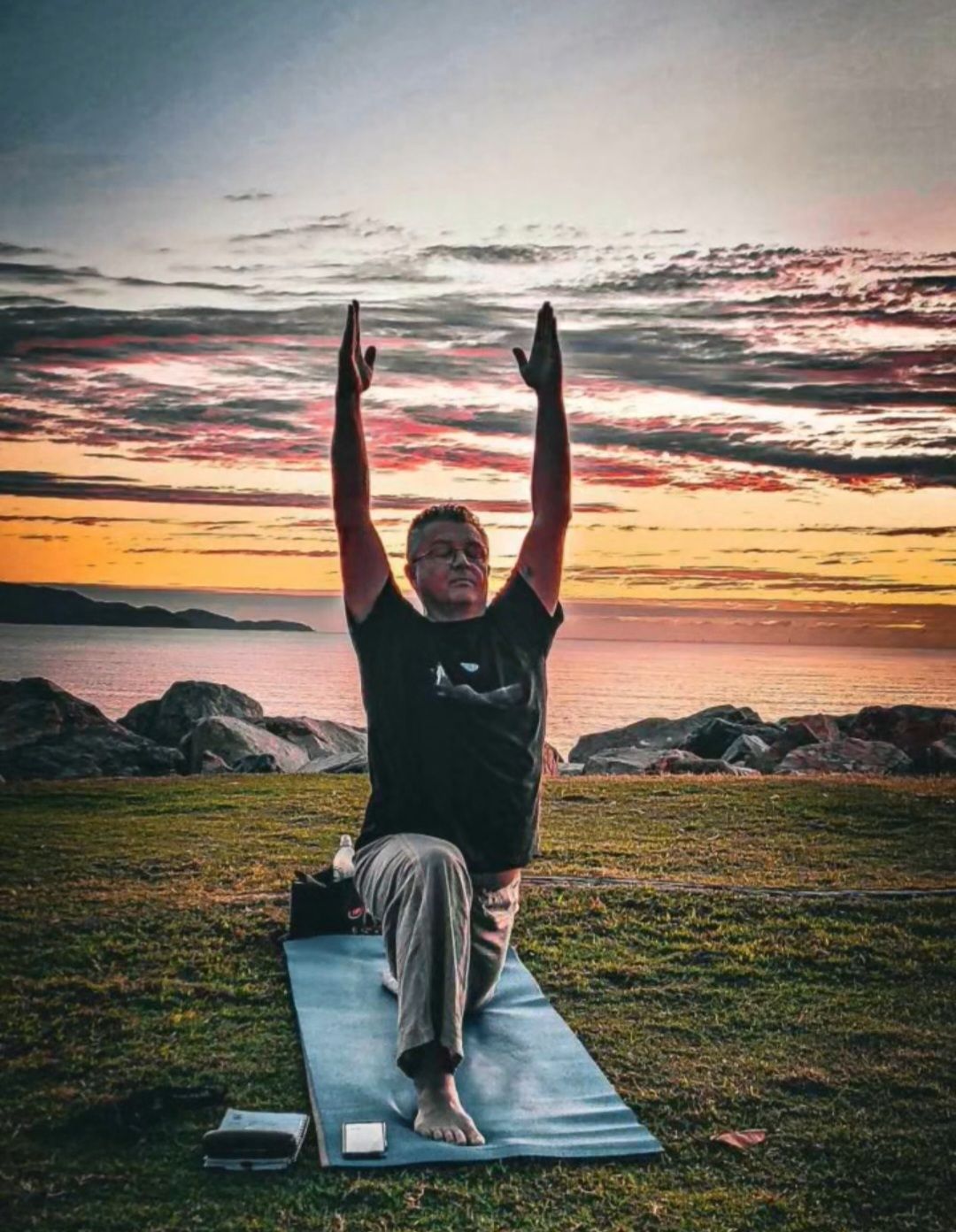 Person in a yoga pose on a mat at sunset by the water, arms raised toward a colourful sky.