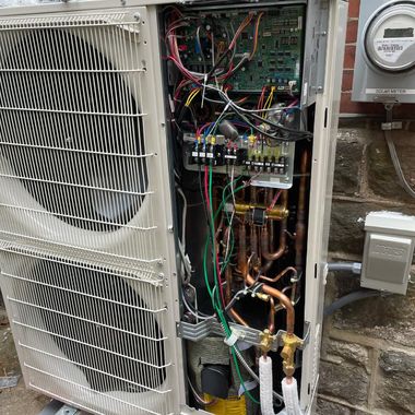 Outdoor HVAC unit with open panel, revealing copper tubing, wiring, and circuit board.