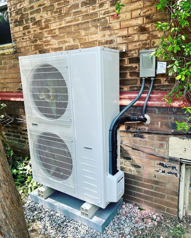 White air conditioning unit on a concrete pad against a brick wall, with electrical box and pipes.