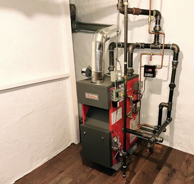 A red and gray furnace with black and copper pipes against a white wall and wood floor.
