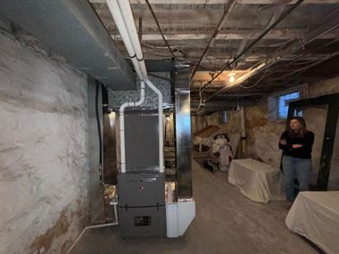 Basement with furnace and ductwork. Person stands nearby, arms crossed. Concrete walls, unfinished ceiling.