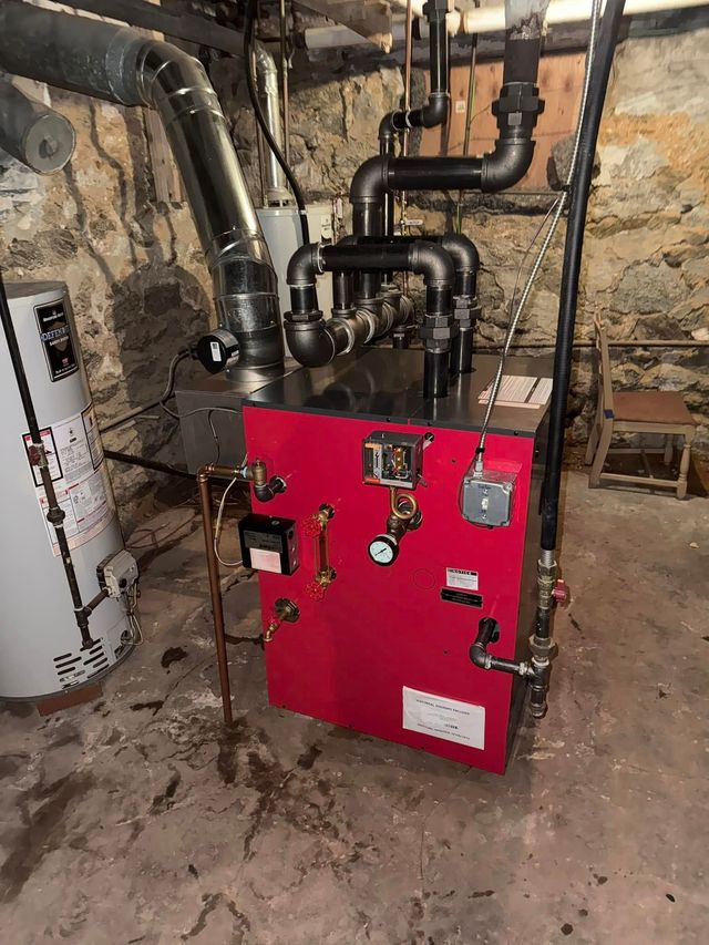A red boiler with black pipes and a water heater in a dimly lit basement.