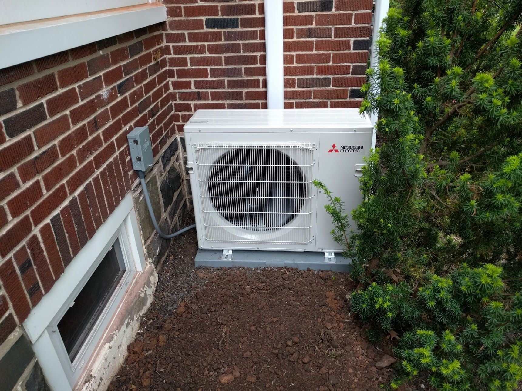 A white Mitsubishi heat pump unit installed outside a brick building next to a bush.