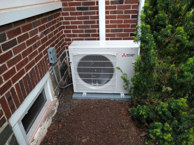 A white Mitsubishi heat pump unit installed outside a brick building next to a bush.