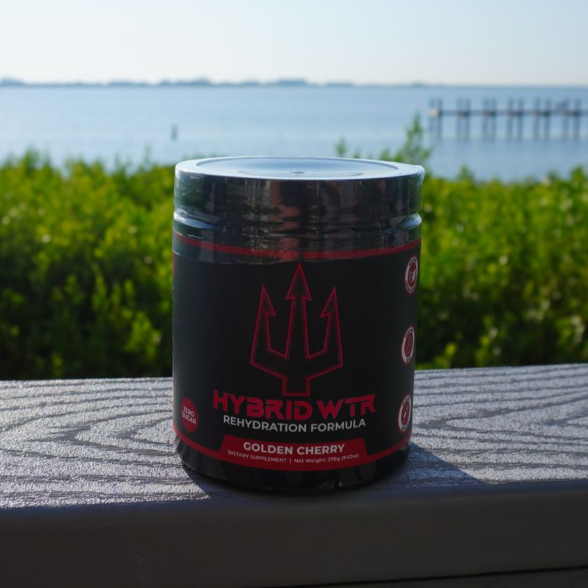 A black tub of "Hybrid WTR" rehydration formula, golden cherry flavor, sits on a wooden deck with a sea backdrop.