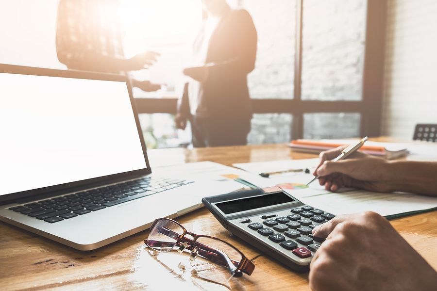 A person is using a calculator and a laptop on a wooden table.