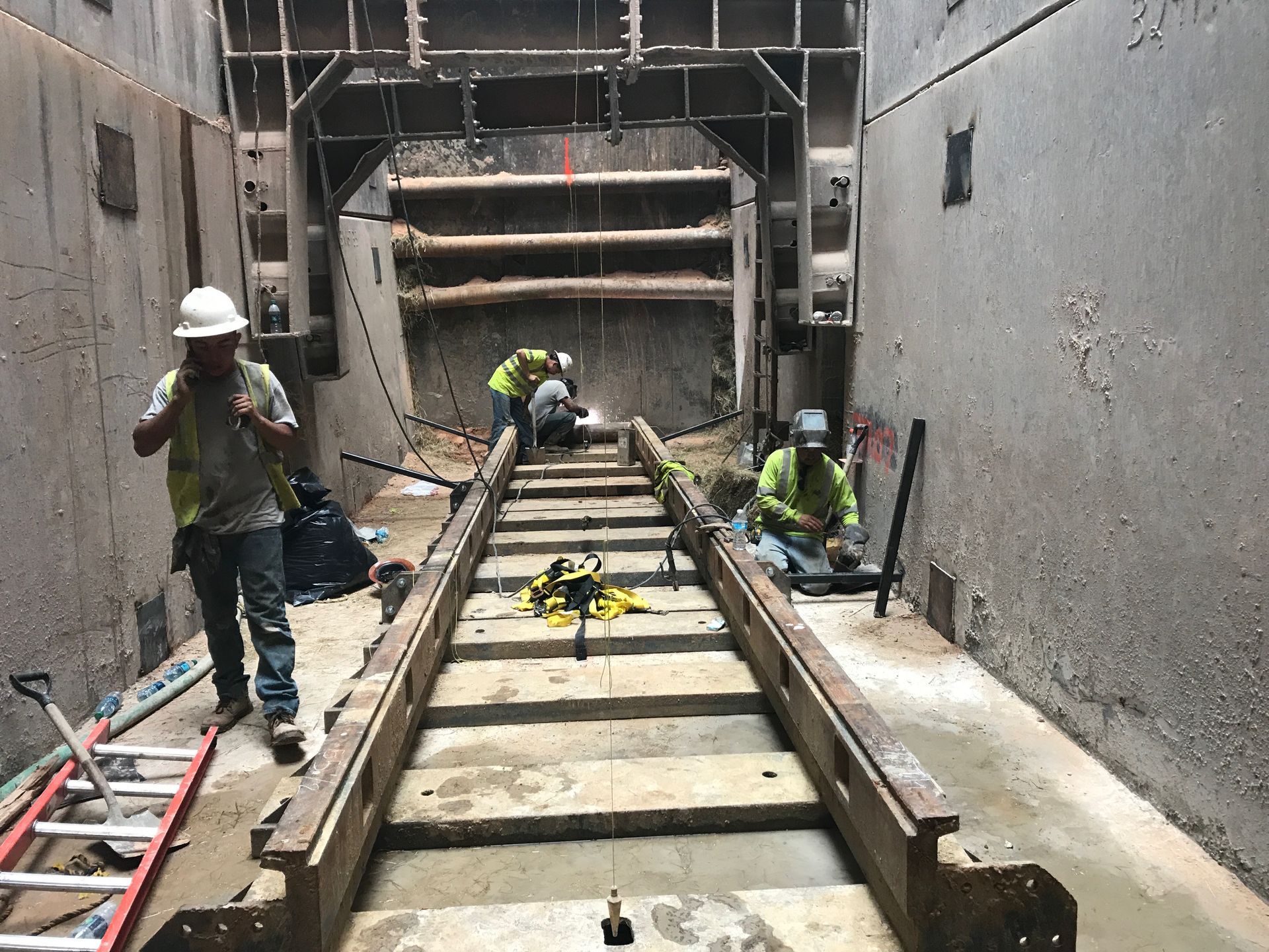 Construction workers in a trench, one on phone, two welding. Heavy machinery frame, concrete walls.