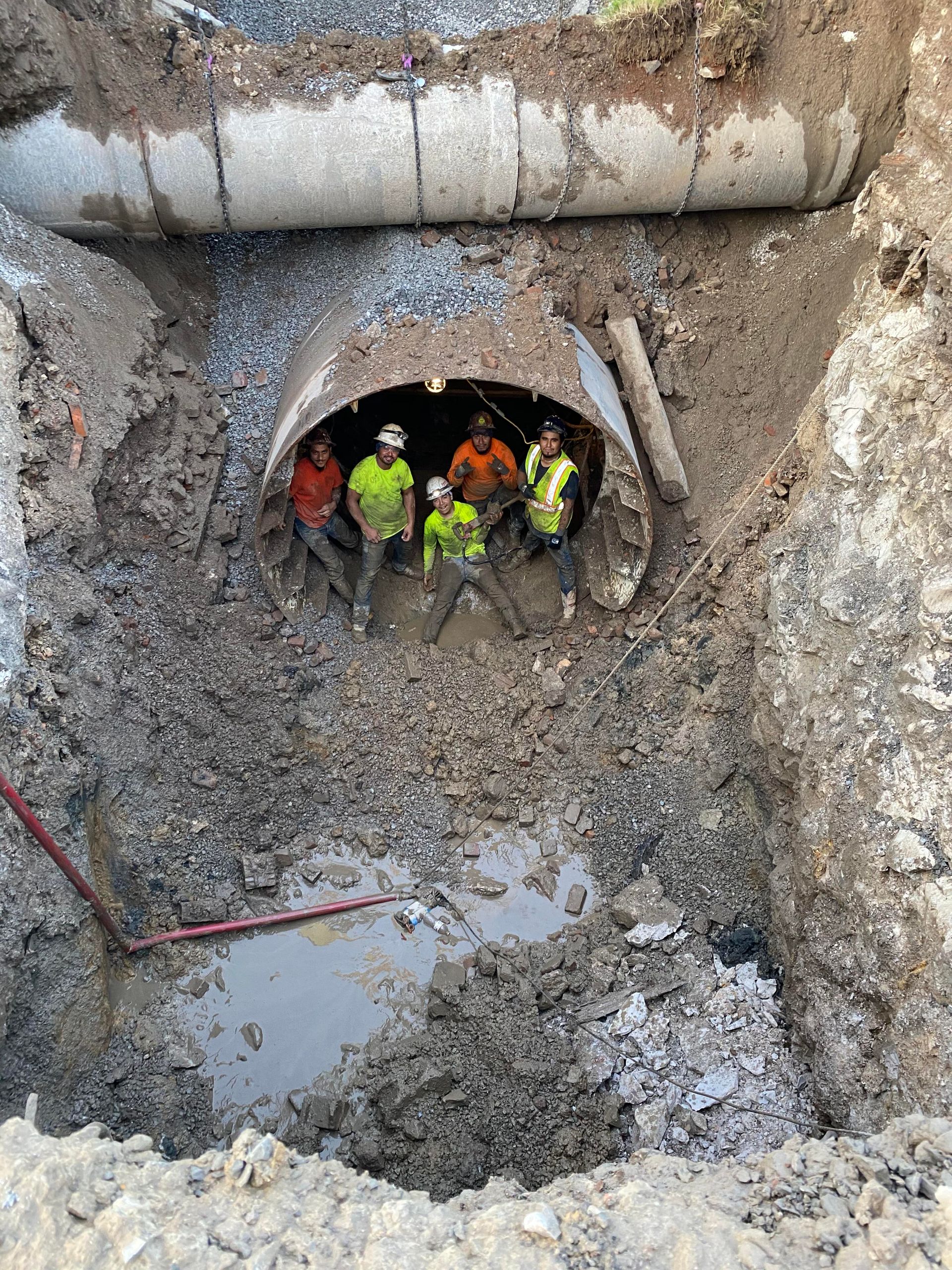 Construction workers inside a dug trench with a large pipe. They wear safety vests and hard hats.