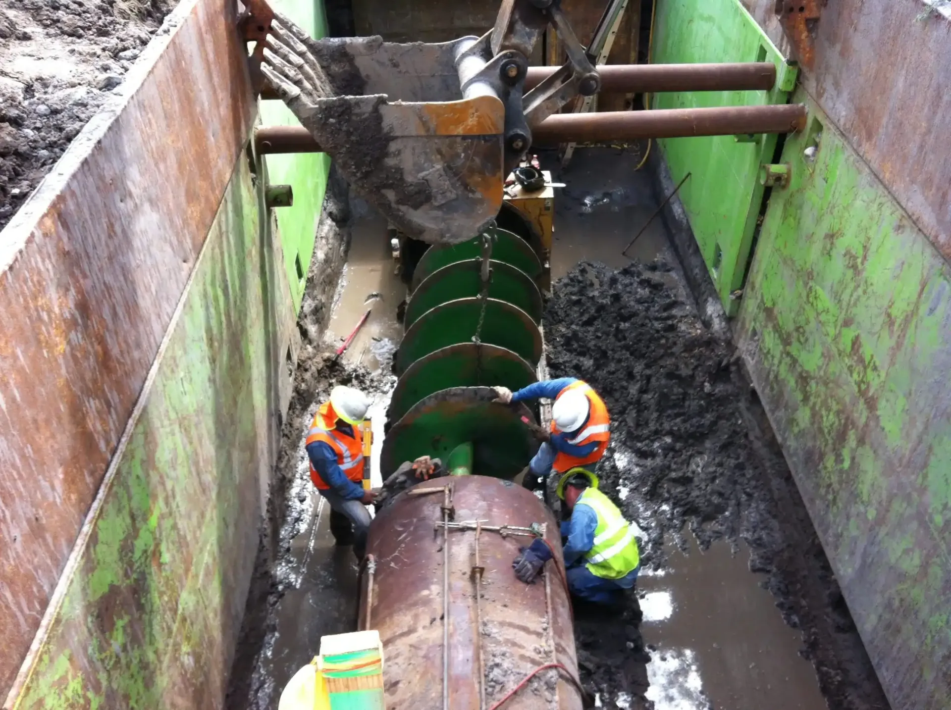 Workers in safety vests and helmets repair a large screw conveyor inside a trench. Mud and metal surround them.