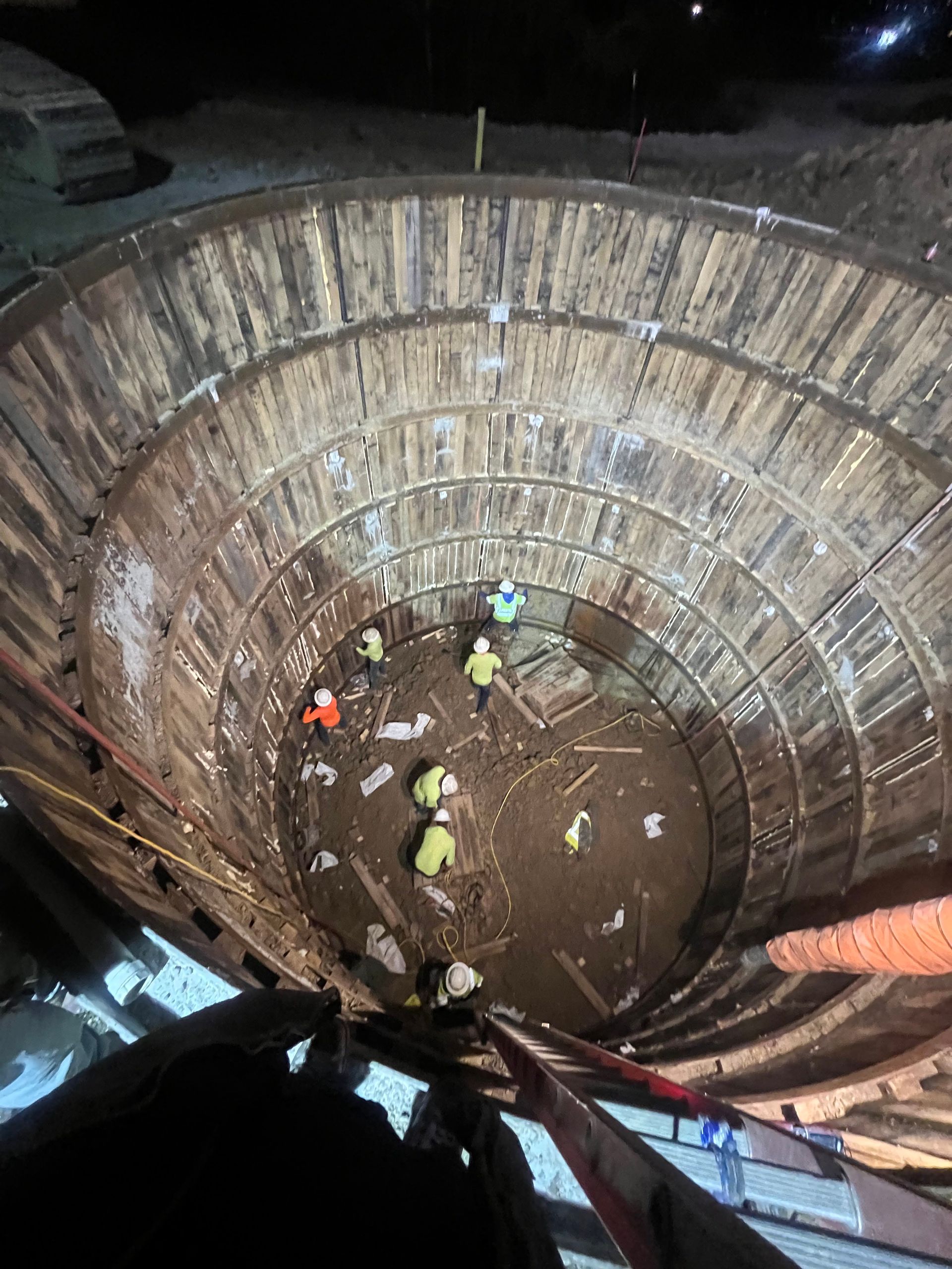 Workers in a deep, circular construction site. Wooden support rings line the walls.