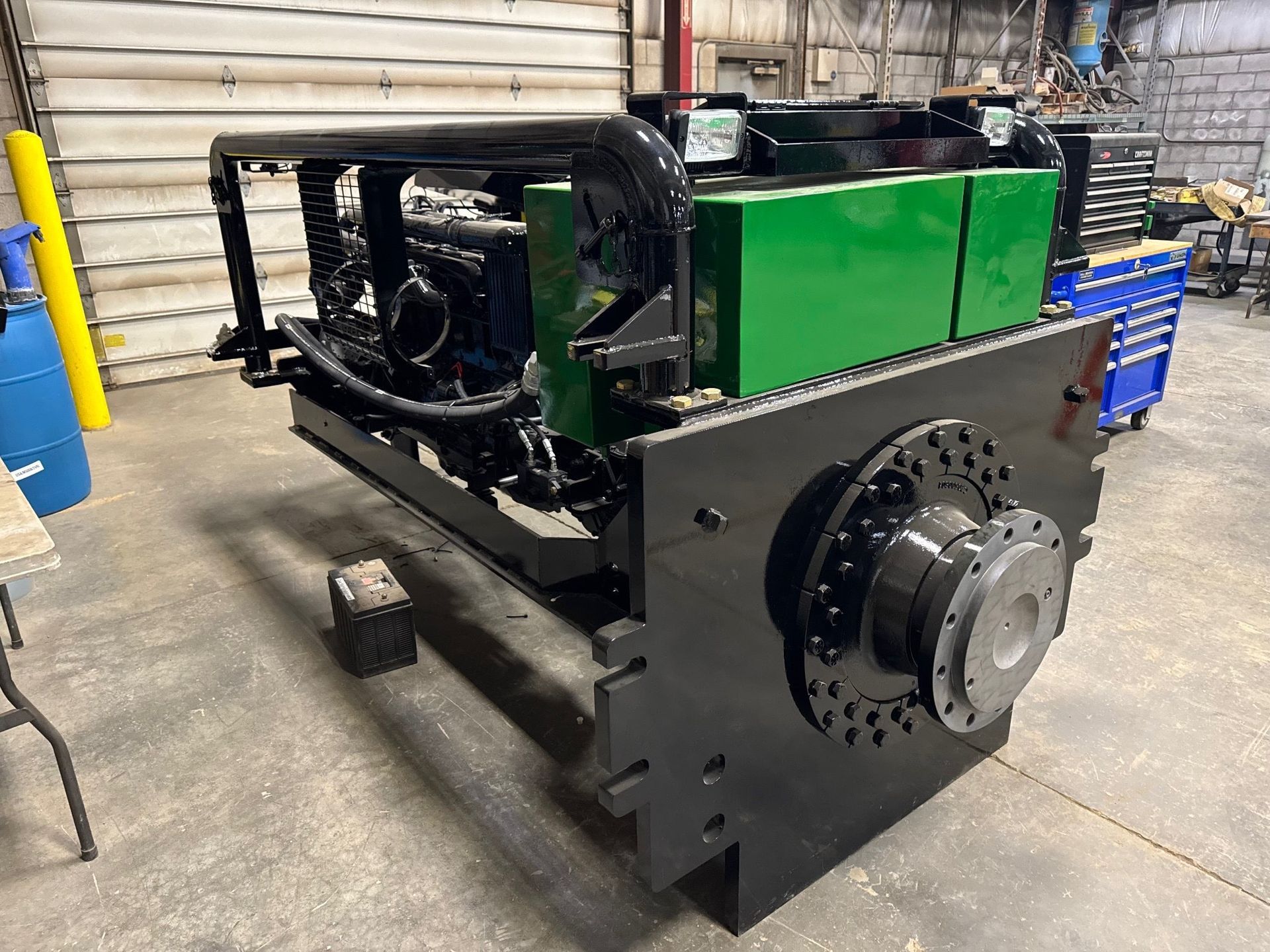 Large black and green industrial machine with circular component attached to a steel frame, set inside a workshop.