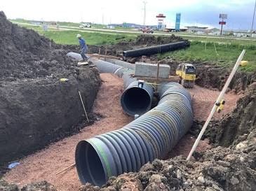 Construction site: corrugated pipes in trench, worker, small excavator, and a road in background.