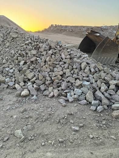 Pile of gray rocks with excavator bucket in a dusty outdoor setting at sunset.