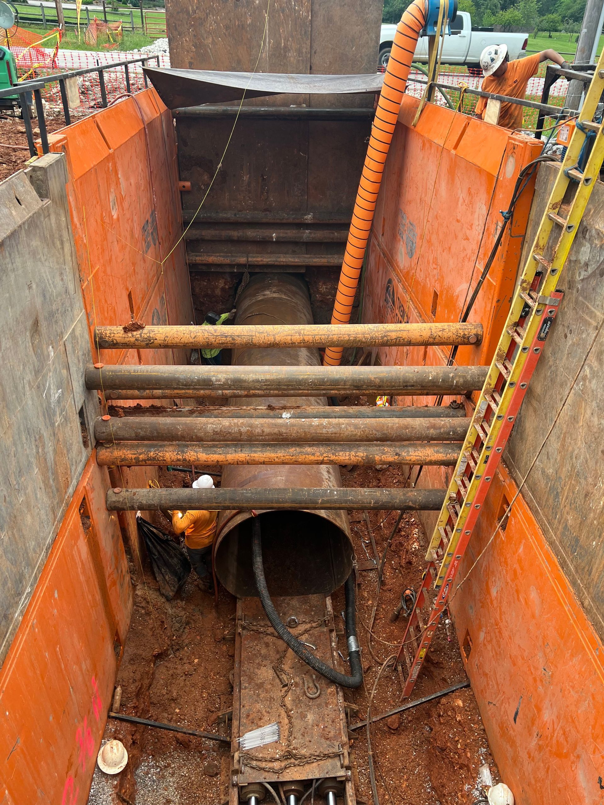 Construction pit with orange walls, a large pipe, and support beams. A ladder stands on the right.