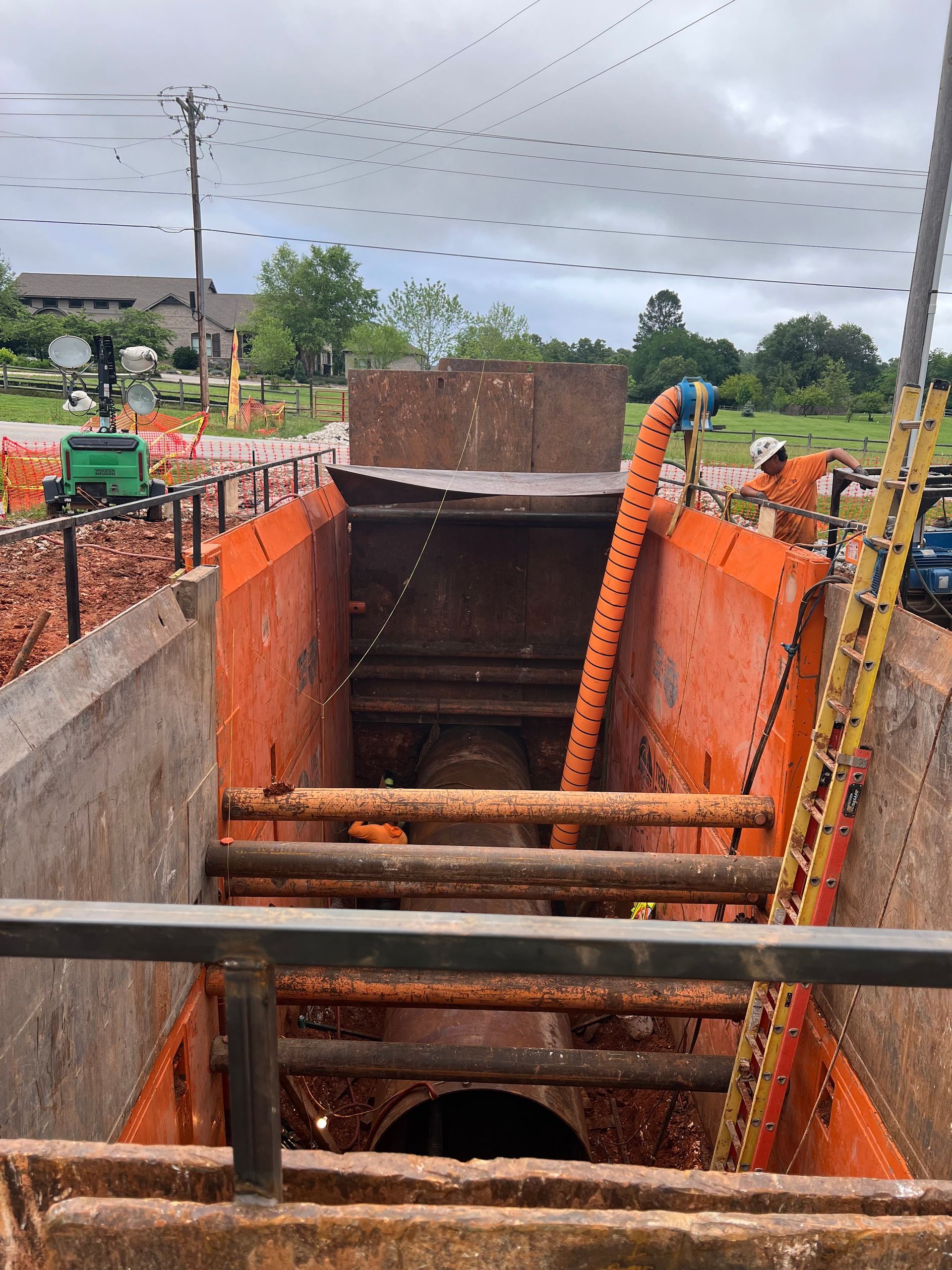 Construction site: Orange-walled trench with pipe, metal supports, and ladder. Machinery visible.