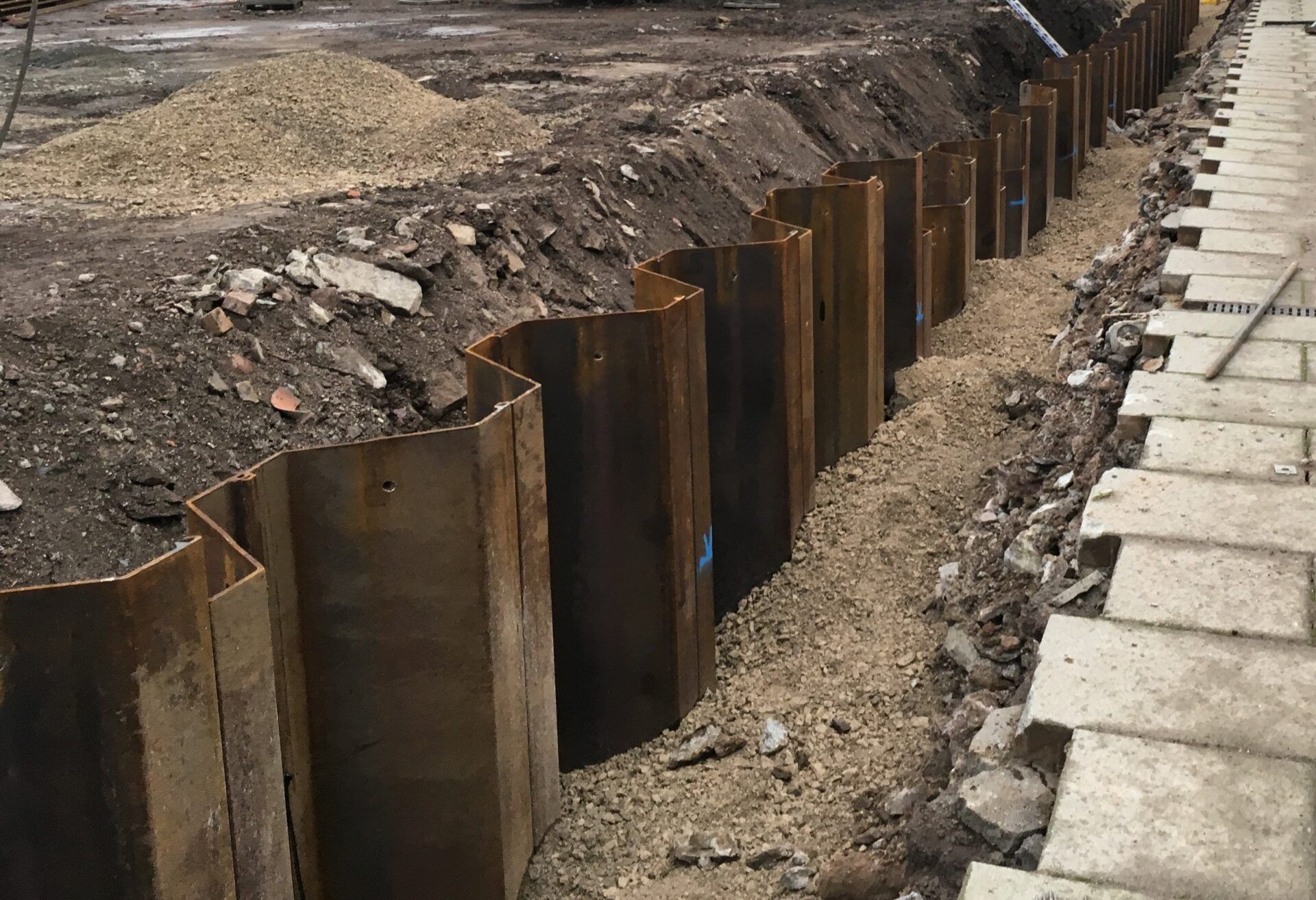 Trench with steel sheet piles, gravel, and construction materials on a city sidewalk.