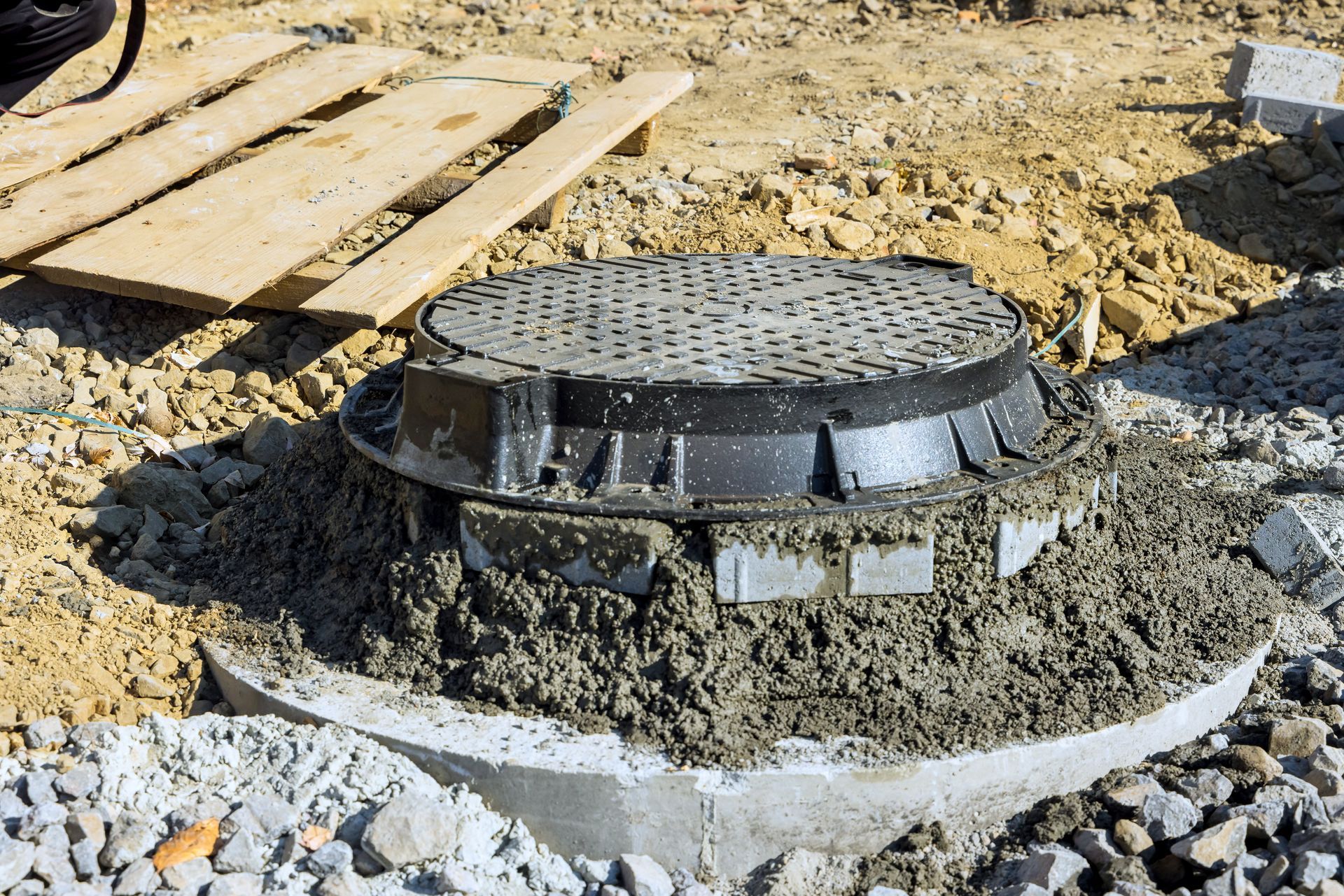 A manhole cover set in concrete during construction. Gravel and wooden boards surround the base.