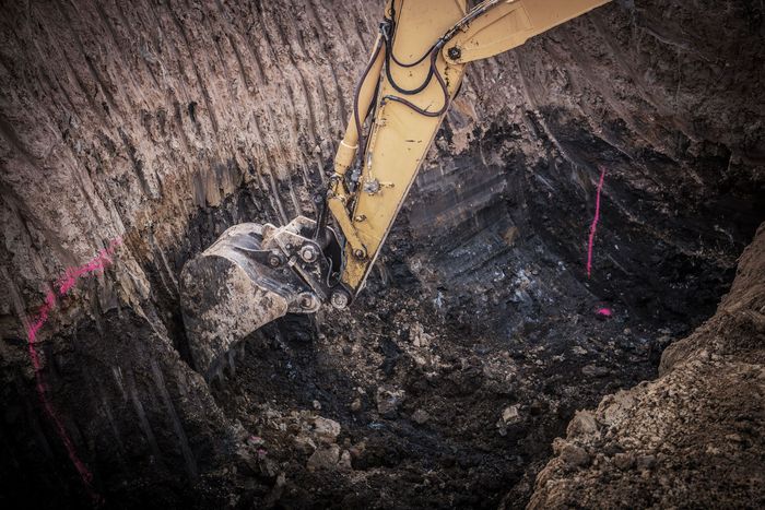 Excavator digging into a dark, layered earth pit; pink spray-painted markers.
