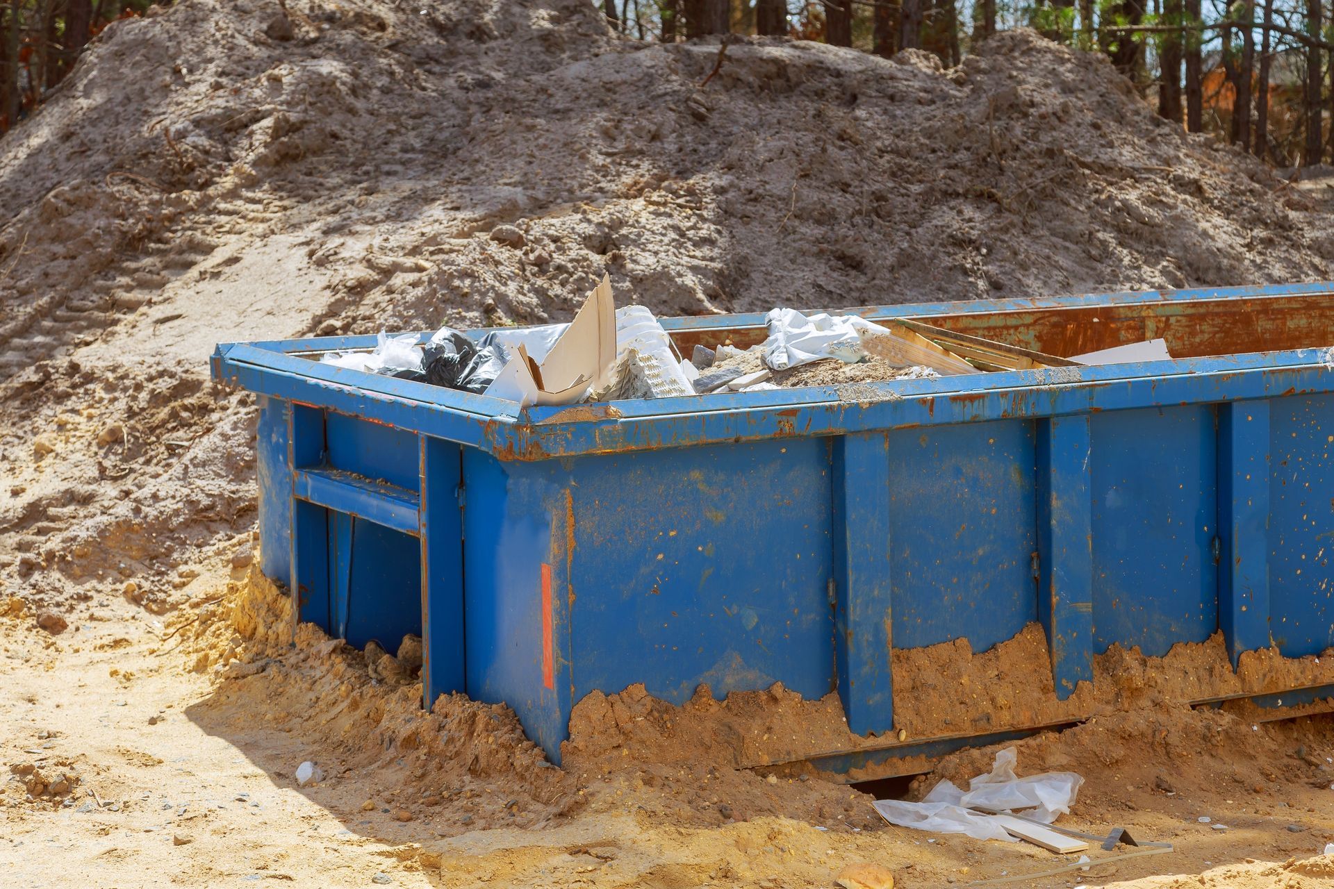 Blue dumpster overflowing with debris at a construction site; surrounded by dirt and a sand pile.