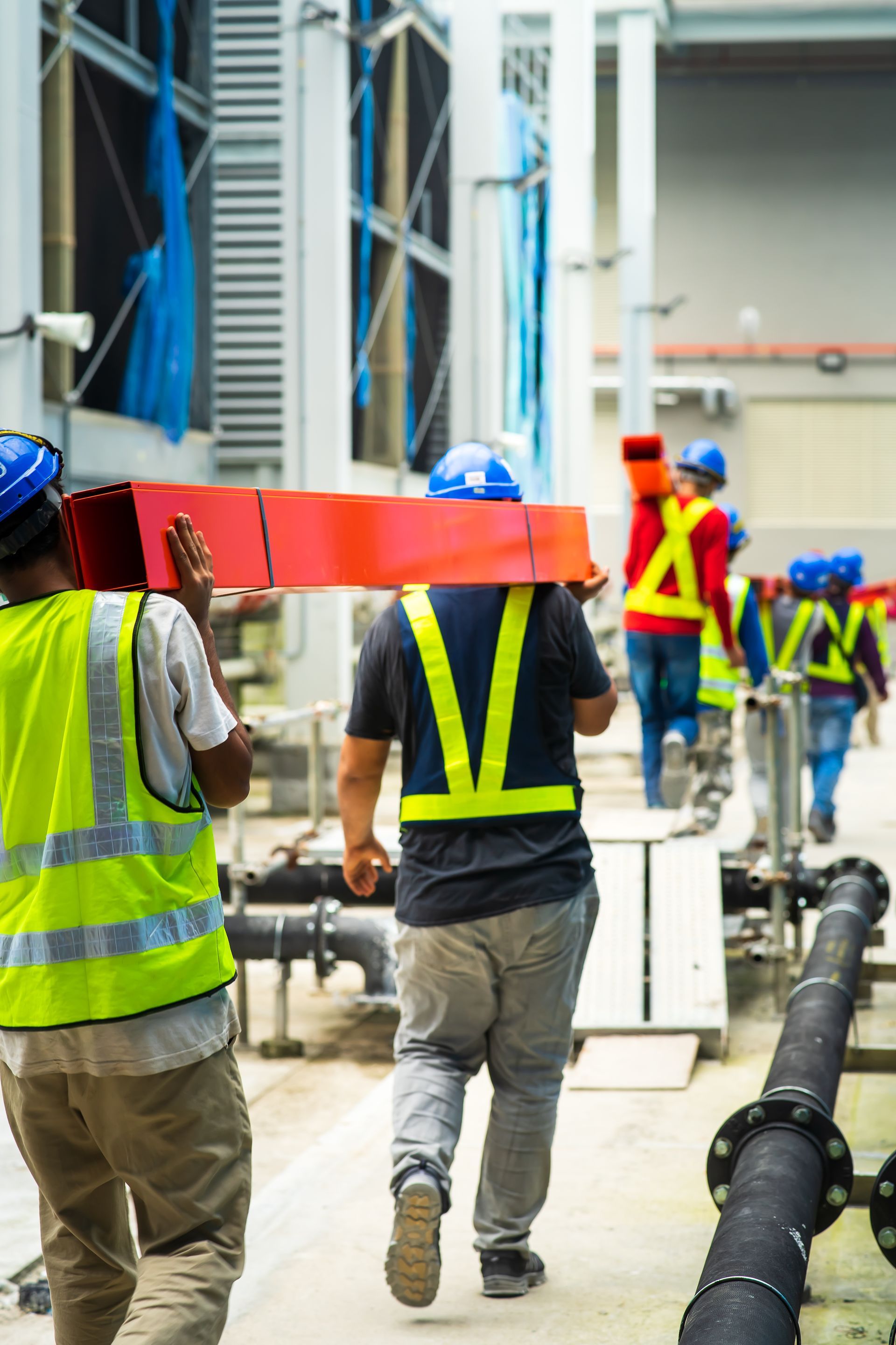 Construction workers carrying materials on a site, wearing safety vests and blue helmets.