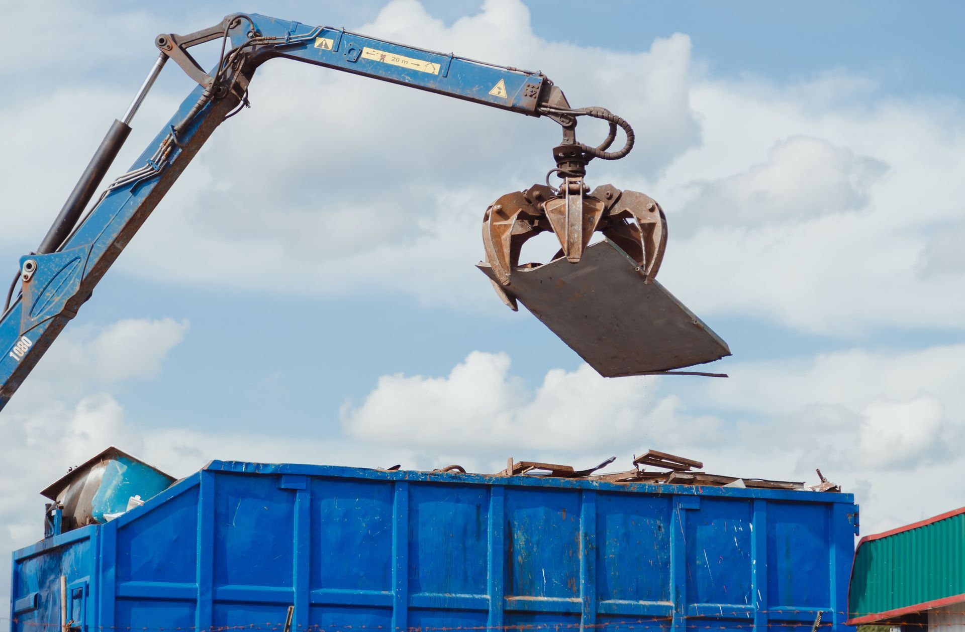 Blue crane loading scrap metal into a blue container under a cloudy sky.