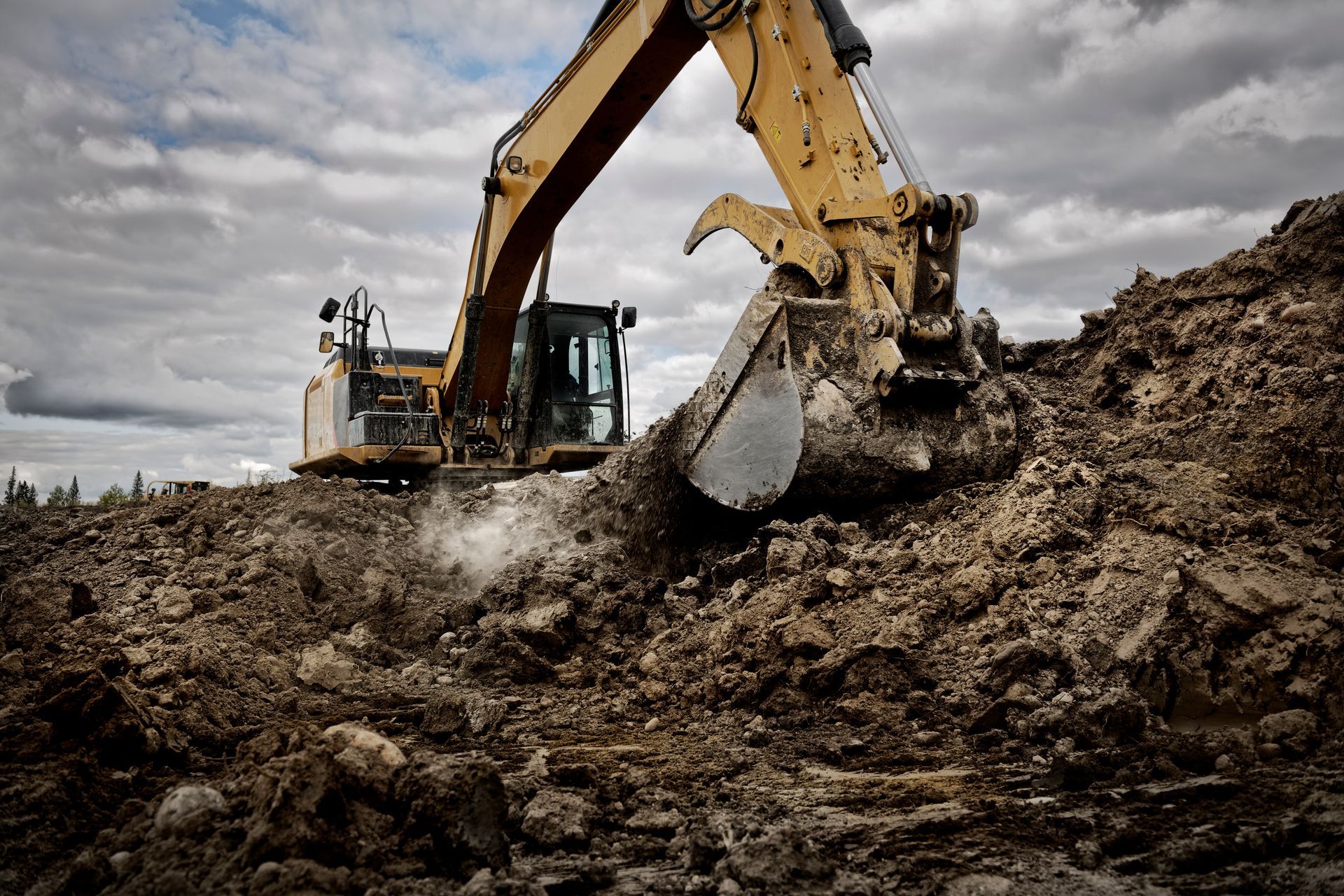 Yellow excavator digging into a pile of dirt under a cloudy sky.