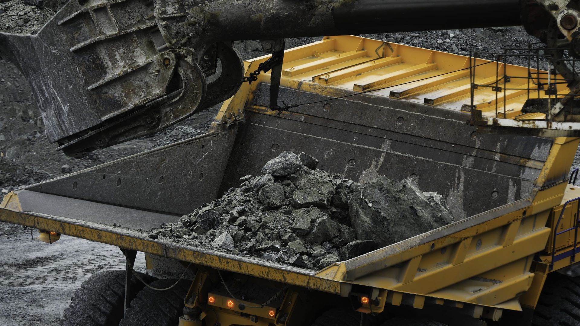 A large yellow dump truck being filled with dark gray rocks by an excavator in a mine.