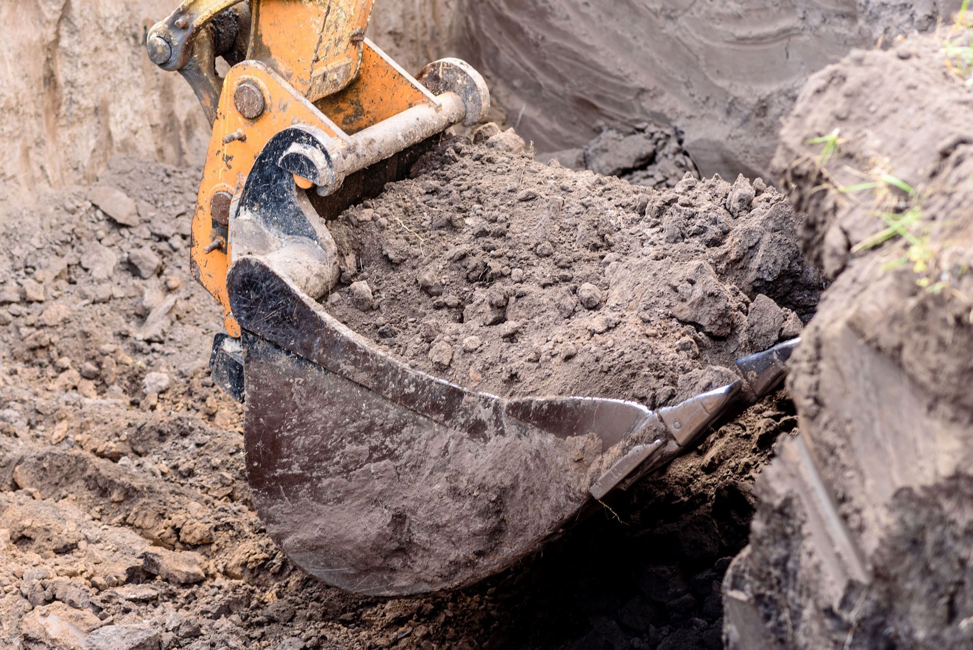 A backhoe bucket scoops dirt from a trench, with exposed soil around it.