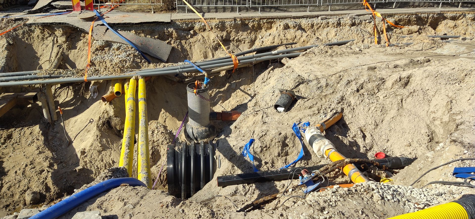 Construction site excavation with yellow and blue pipes, and black ribbed pipe, and dirt.