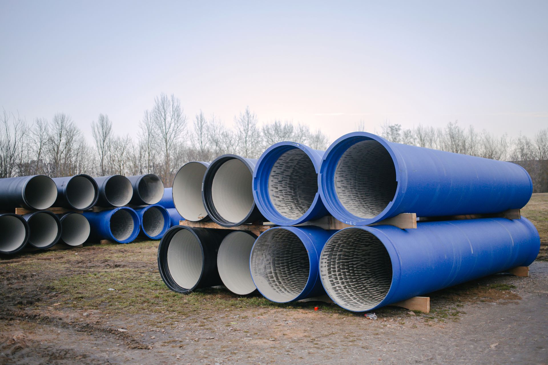 Blue and gray pipes stacked on a grassy area, trees in the background.
