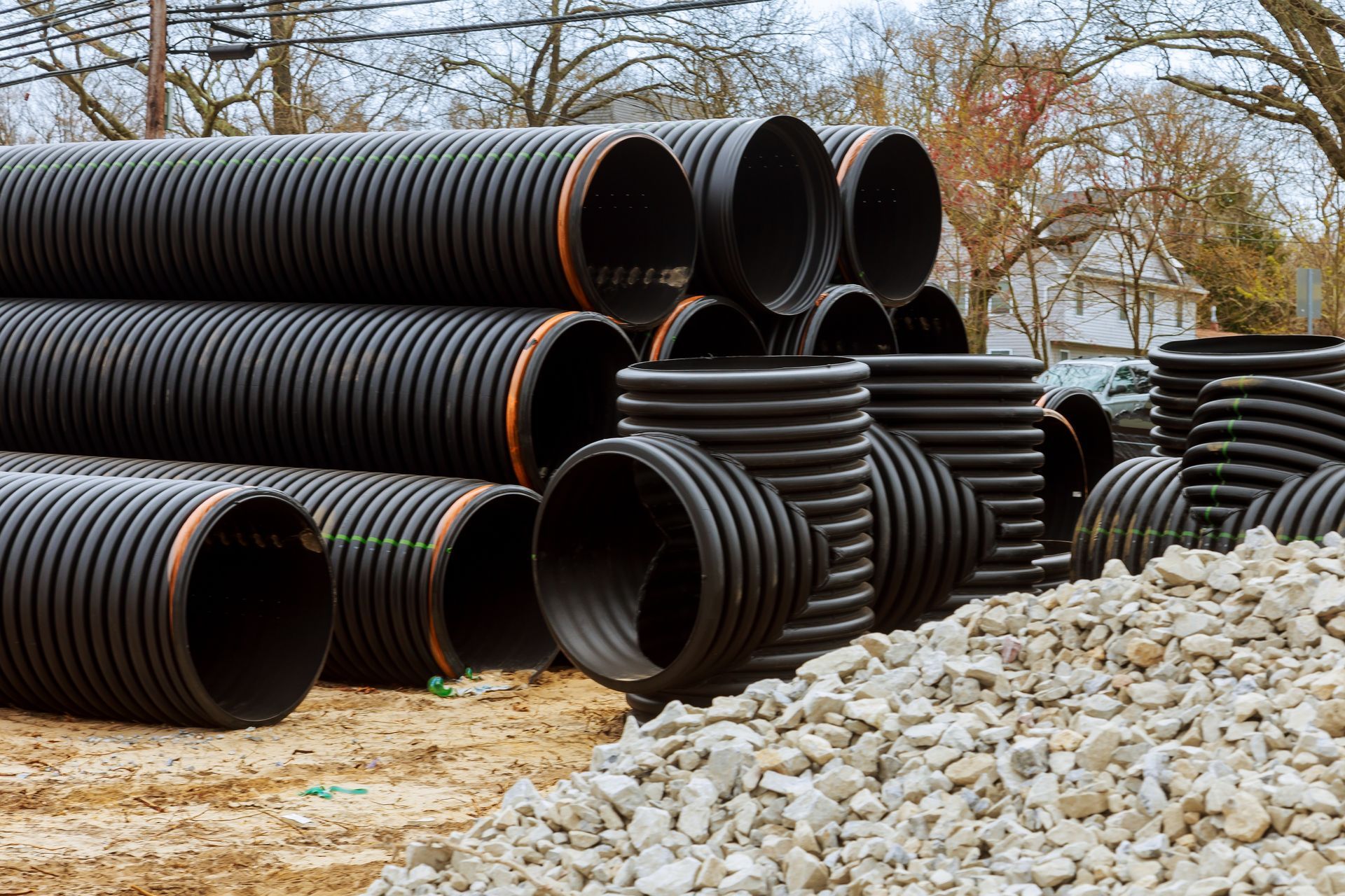 Black corrugated drainage pipes stacked near gravel pile outdoors.
