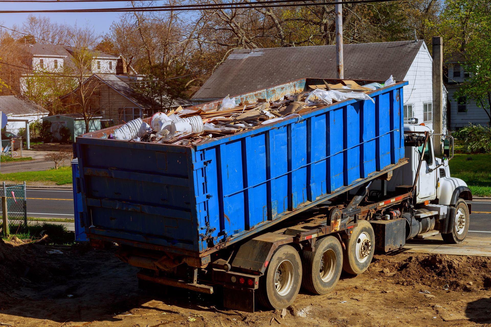 Blue dumpster being lifted by a truck, filled with debris, in front of houses.