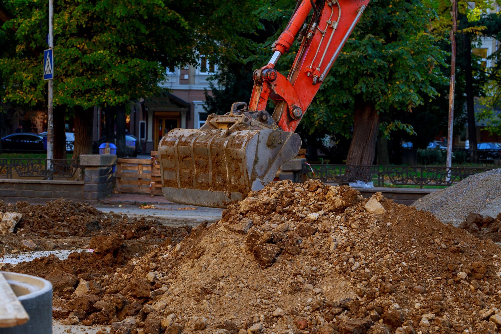 An orange excavator bucket scoops dirt from a construction site next to a pile of soil in front of trees.