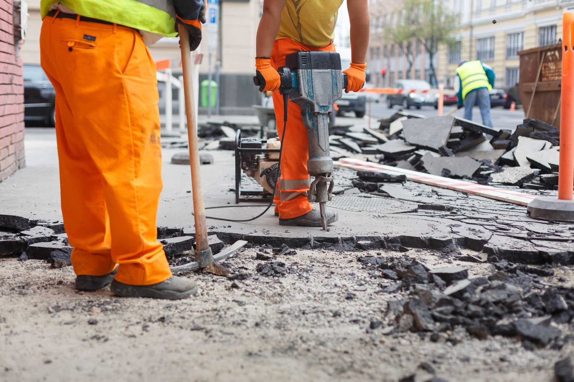 Construction workers in orange pants break asphalt with a jackhammer and tool in city.