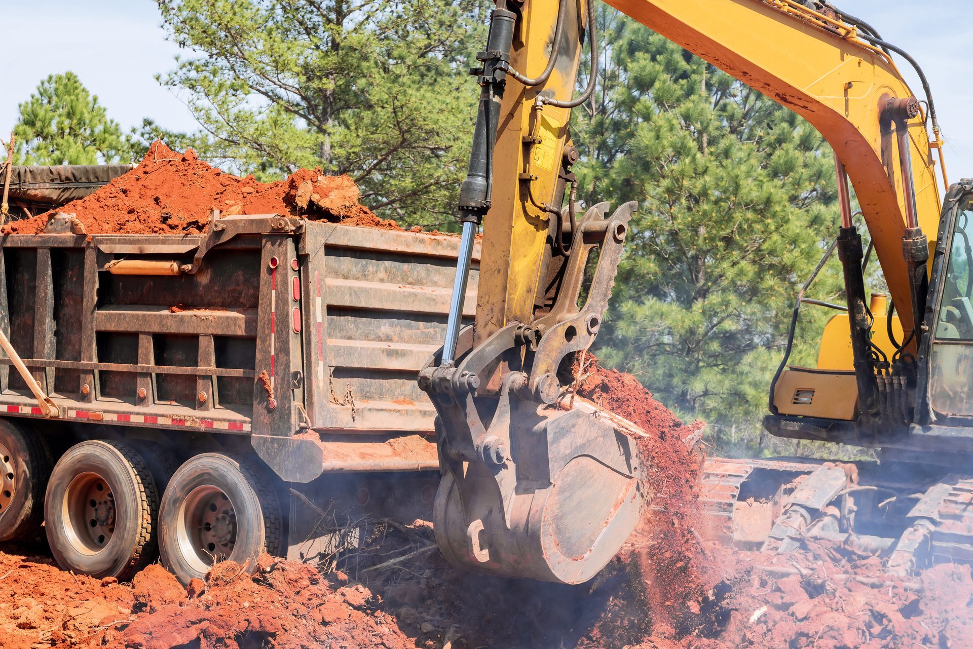 An excavator loading reddish dirt into a dump truck at a construction site.