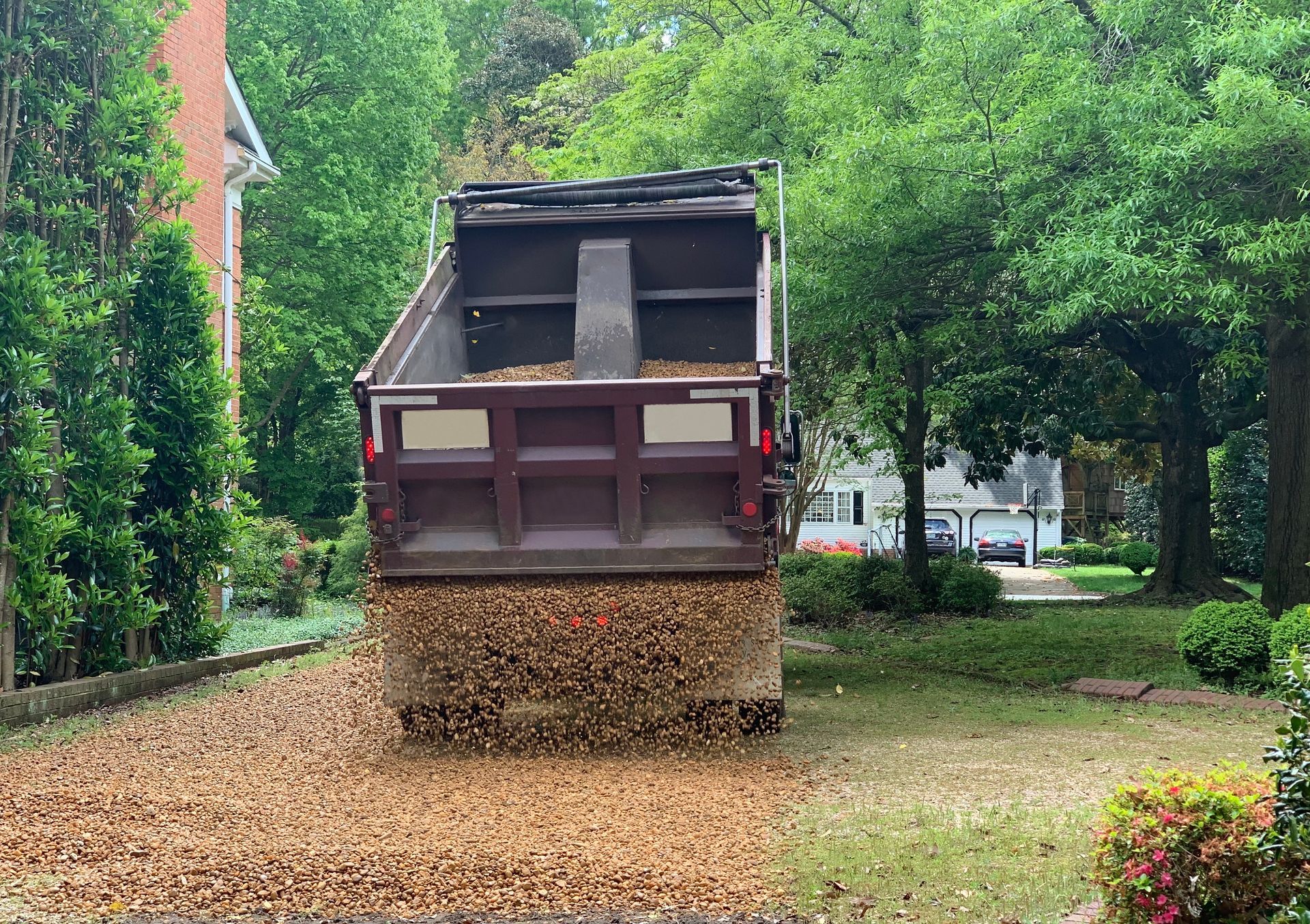 Dump truck unloading mulch onto a driveway. Brown mulch, green trees, and a house in the background.