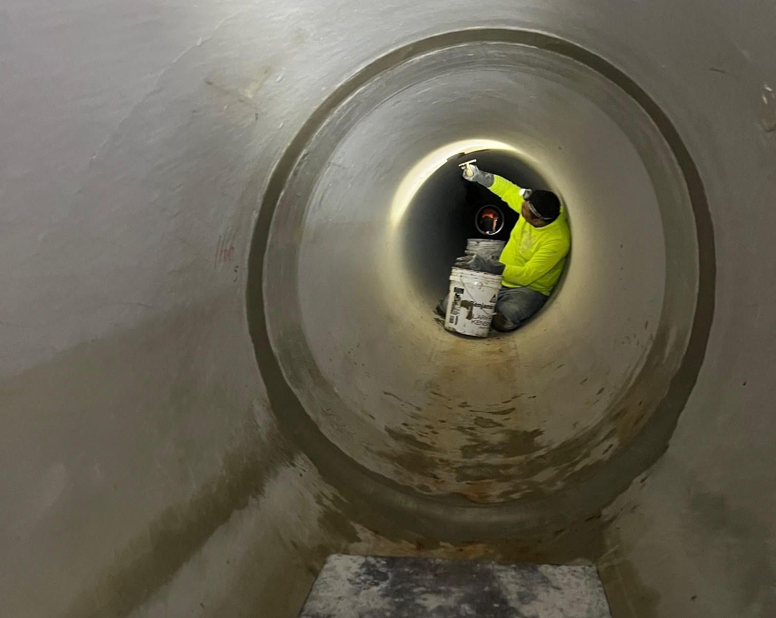 Person in yellow jacket repairing the inside of a large, circular concrete pipe.