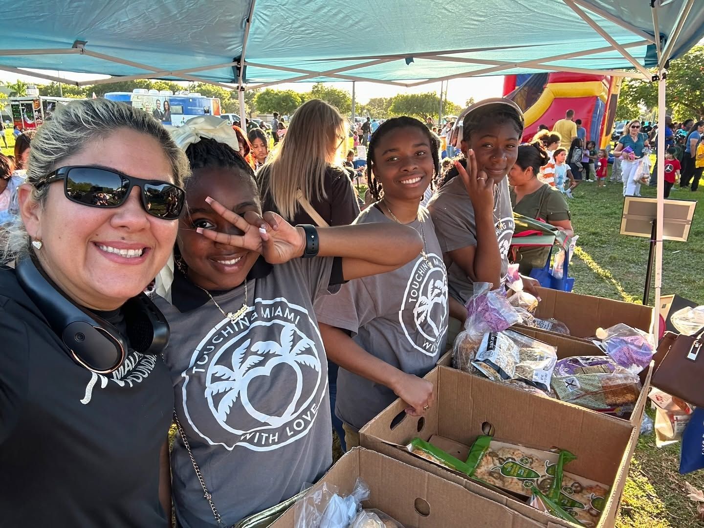 A woman and three girls smile at a table selling items at an outdoor event. They are under a blue tent.