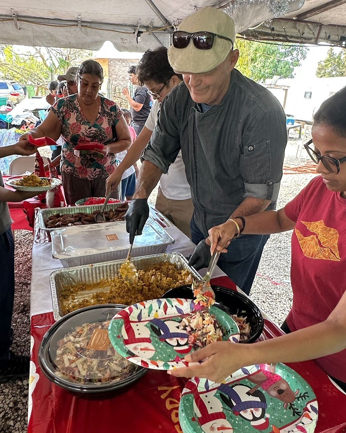 People being served food from a buffet table outdoors. A man in a cap and gloves serves from a tray. Others wait with plates.