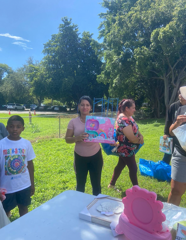 A woman holds a toy in a park, surrounded by people and greenery, possibly at an event. A child stands nearby.