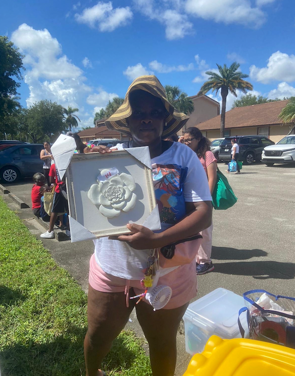 Person in straw hat holding a framed floral wall decoration outdoors, possibly moving. Cars, people, and buildings are visible in the background.