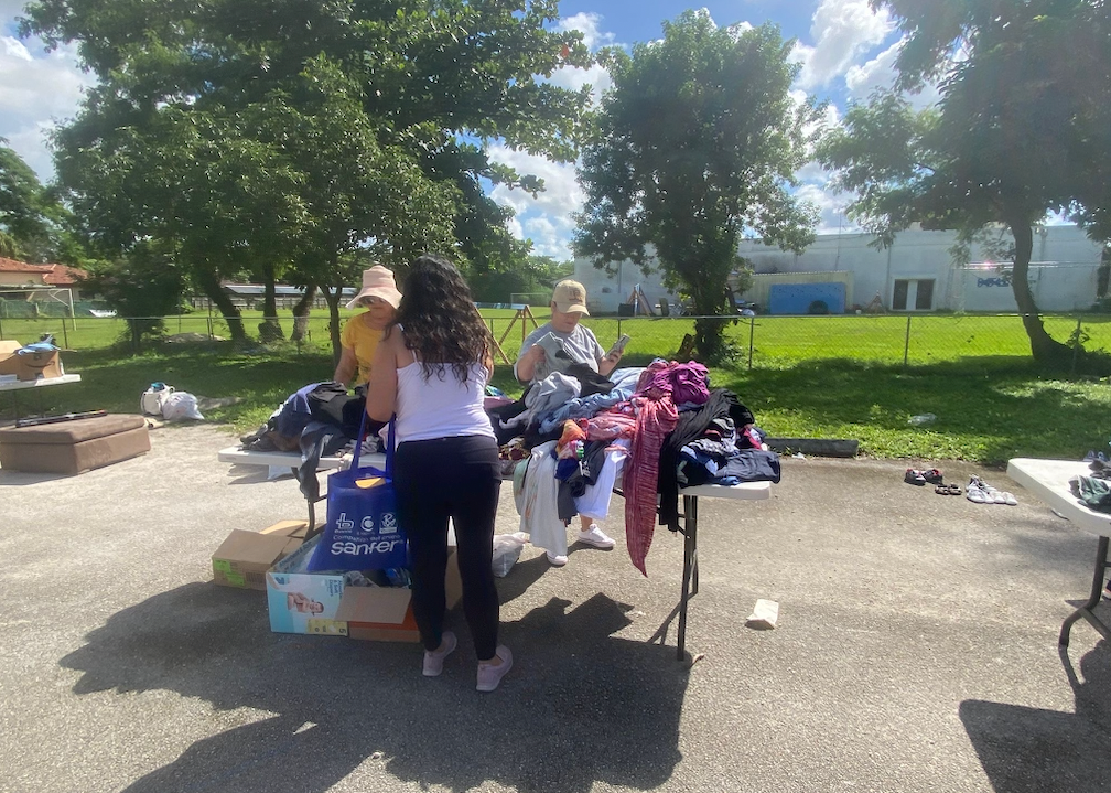 People browsing clothes on tables at an outdoor sale on a sunny day. Trees and a building are in the background.