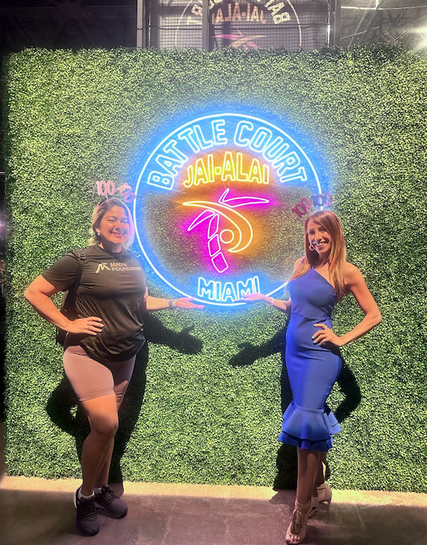 Two women pose with arms outstretched next to a neon sign for 