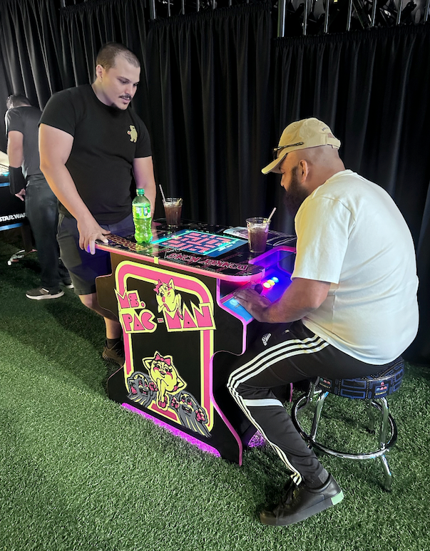 Two men at a custom arcade machine. One plays while seated, the other watches. Bright pink, black, and purple arcade design.