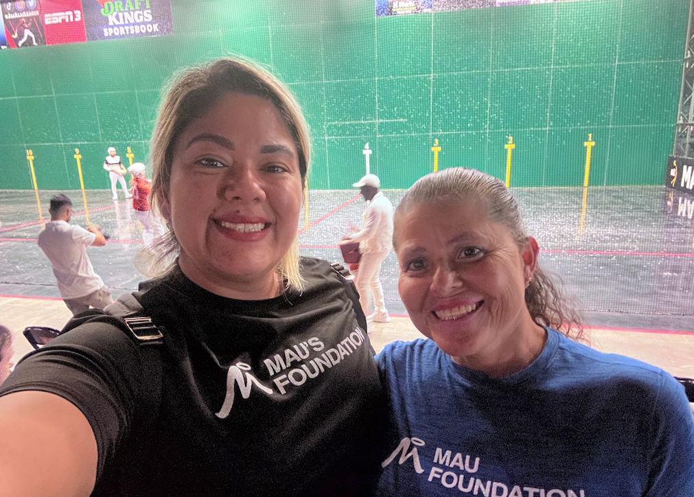 Two smiling women take a selfie at a jai alai game. One wears a black shirt with a logo; the other, a blue shirt. Players are in the background.