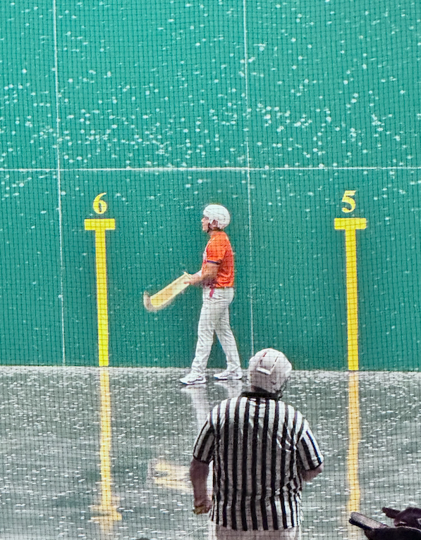 A handball player in an orange shirt prepares to hit the ball. A referee watches in the foreground, with the fronton wall as a backdrop.